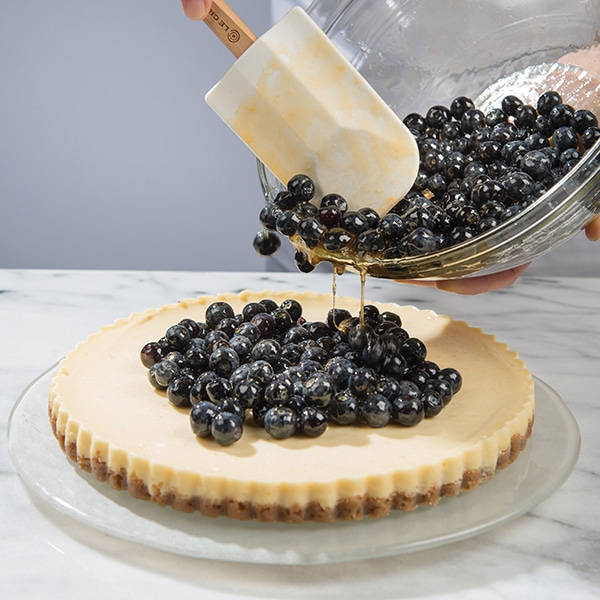 Blueberry compote being poured on top of a fluted lemon cheesecake